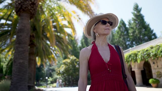 Mature woman smiles and looks around while touring a winery farm in Napa with sunshine and palm trees in the background