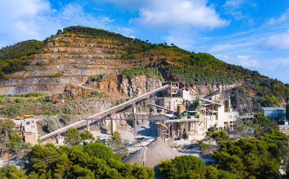 Aerial view of the Cava di Colle Reciso quarry featuring industrial machinery, conveyor belts, and gravel piles set against a terraced hillside in Portoferraio, Toscana, Italy.