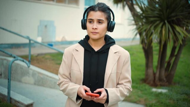 Young Latina woman walking through an urban area while listening to music with headphones and using her smartphone, showing a modern connected lifestyle.
