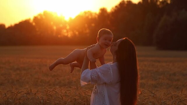 A happy mother lifts her seven-month-old son above her head in a wheat field in the rays of the setting sun and kisses him tenderly