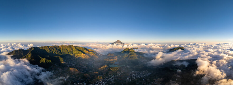 Aerial view of the Dieng plateau featuring volcanic mountains, lush green hills, and small villages nestled among clouds under a clear blue sky in Kejajar, Central Java, Indonesia.