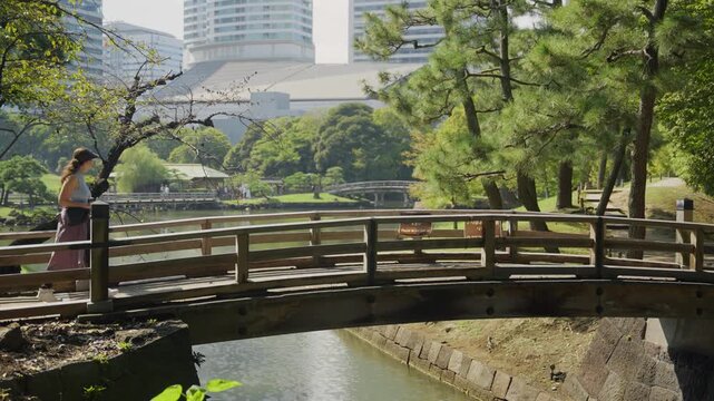 Young woman walking across a wooden bridge over a moat in the Hamarikyu Gardens in Tokyo, Japan, with city buildings visible in the background on a sunny day in late spring