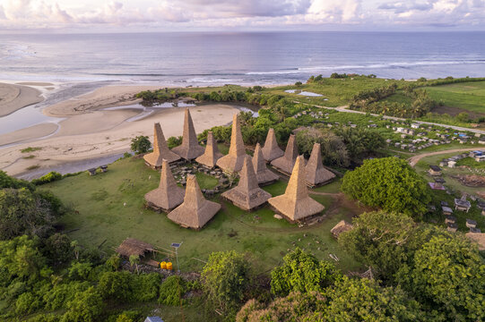 Aerial view of Kampung Adat Ratenggaro featuring traditional high-roofed houses situated on a grassy hill overlooking the beach and ocean in Kodi Bangedo, Sumba, Indonesia.