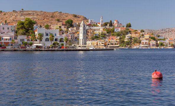 View of the waterfront with the historic clock tower and colorful hillside houses in Symi Harbor, Dodecanese, Greece.