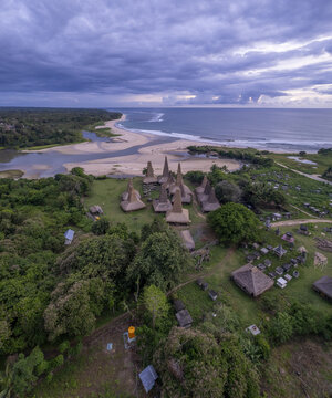 Aerial view of Kampung Adat Ratenggaro with its traditional high-roofed houses, lush green surroundings, and nearby sandy beach along the coastline in Kodi Bangedo, Sumba, Indonesia.