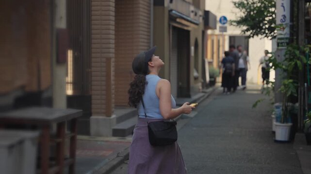 Young tourist woman with a cap and curly hair walking through a narrow, traditional alley in Tokyo, curiously looking around at the historic architecture and soaking in the local atmosphere