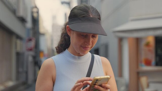 Young Caucasian woman with a sun visor texting or checking her smartphone while standing in a narrow urban alley in Tokyo, Japan, navigating city or communicating with friends during her vacation