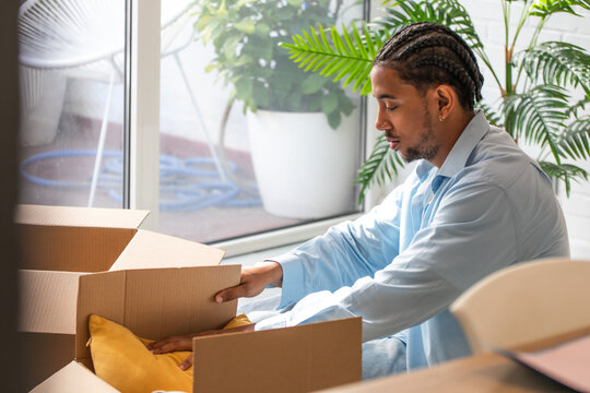 Smiling young man with braided hair unpacking cardboard boxes while moving into a new home, sitting among