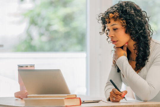 Young woman with curly hair working from home, writing notes while using a laptop at a wooden table, focused on studying or remote work in a bright, modern living room