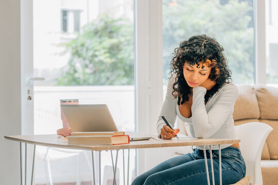 Young woman with curly hair working from home, writing notes while using a laptop at a wooden table, focused on studying or remote work in a bright, modern living room
