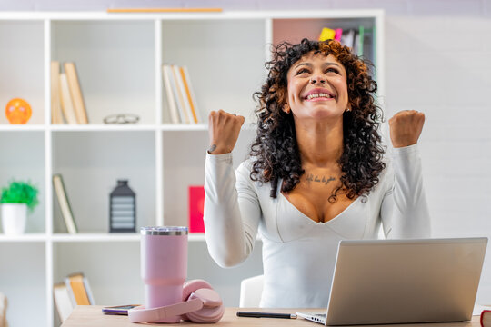 Excited young woman with curly hair celebrating success at her desk, raising both fists in triumph while working on a laptop in a modern home office
