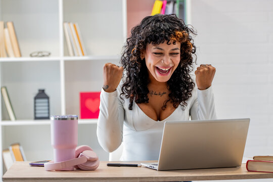 Excited young woman with curly hair celebrating success at her desk, raising both fists in triumph while working on a laptop in a modern home office
