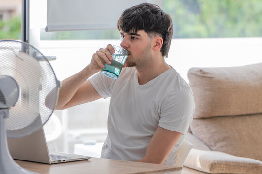 Young man cooling off with an electric fan while drinking a glass of water at his desk during a summer heatwave, sitting in front of an open laptop at home