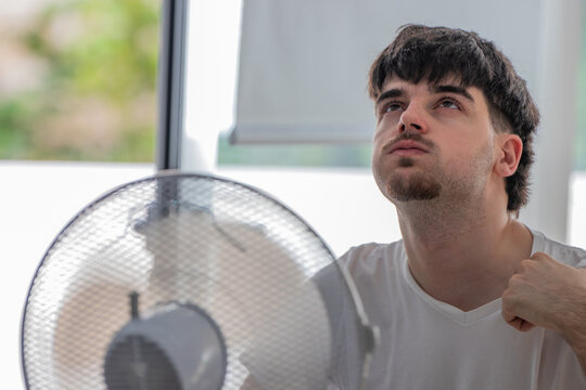 Young man sitting by a pedestal fan, tilting his head back and closing his eyes to feel the cool breeze while trying to stay cool during a hot summer day indoors