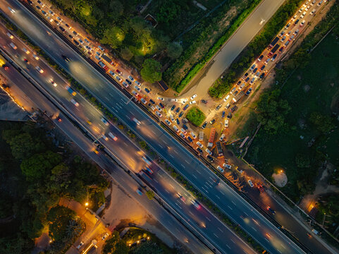 Aerial view of a busy highway interchange at night with glowing vehicle headlights and taillights creating light trails on the asphalt roads surrounded by urban greenery in Gurgaon, Haryana, India.