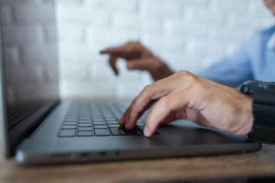 Side close-up of a man's hands typing on a grey laptop keyboard. One finger is pressing a key, while the other is blurred and pointing in the background against a white brick wall.