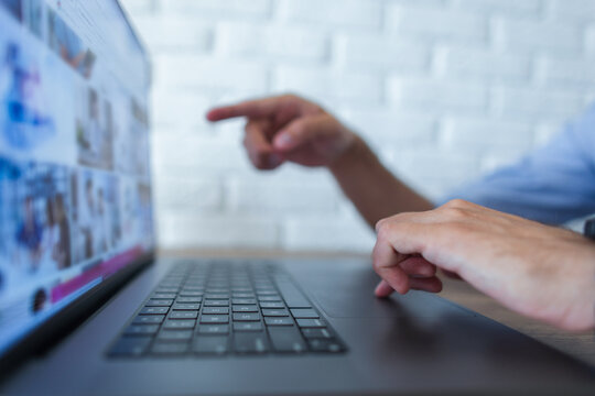 Close-up of a person's hands using a modern laptop on a wooden desk, pointing a finger at the screen while browsing a web page or photo gallery. Shallow depth of field, white brick wall background.