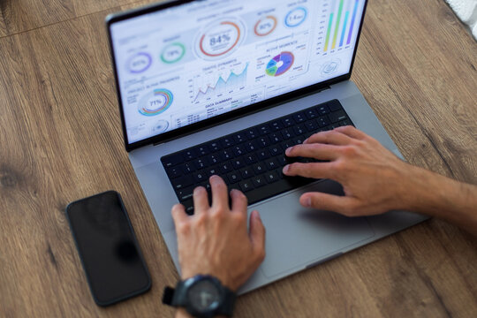 Top view of a man's hands typing on a laptop keyboard while analyzing a financial dashboard with charts and graphs on the screen. A smartphone lies nearby on the wooden surface.