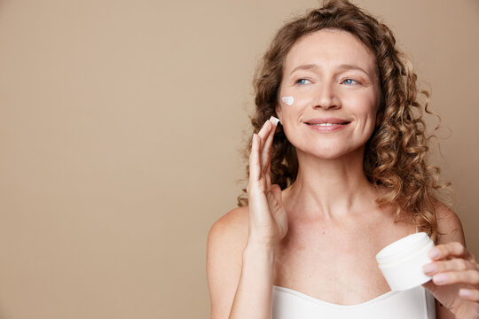 Authentic middle aged woman applying white cream on her cheek, natural skin, no retouching, soft studio lighting, warm mood, self care and skincare concept.