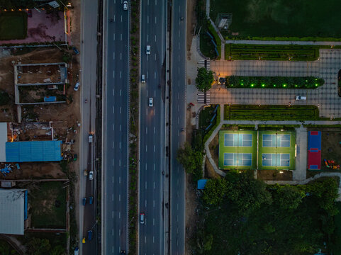 Aerial view of a highway with moving cars at dusk, adjacent to illuminated tennis courts and landscaped park areas in Gurgaon, Haryana, India.