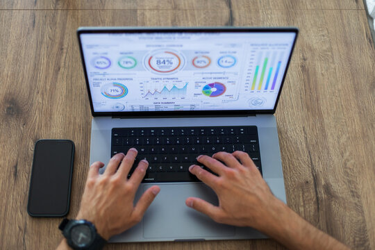 Overhead view of a man's hands typing on a laptop displaying a financial analytics dashboard on a wooden desk. A smartphone mockup lies nearby. Perfect for business and tech concepts.