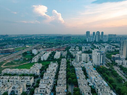 Aerial view of residential neighborhoods with rows of houses, green trees, and modern high-rise buildings in the distance under a soft sunset sky in Gurgaon, Haryana, India.