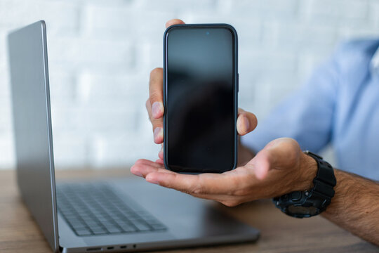 Close-up of a man holding a black smartphone with a blank mockup screen in his hand next to a laptop on a wooden desk. White brick wall blurred background. Modern workspace concept.