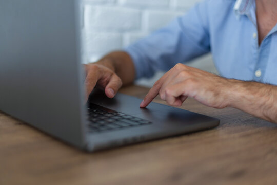 Close-up side view of a man's hands using a modern laptop on a wooden desk. One finger is touching the trackpad while navigating. Bright office with a white brick wall blurred background.