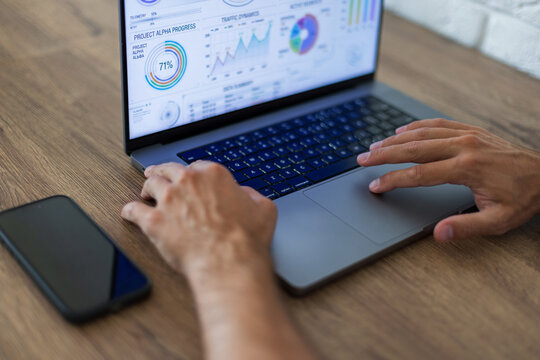 Close-up side view of a businessman's hands typing on a laptop with financial analytics on screen. Smartphone on a wooden desk, blurred white brick wall background. Modern workspace concept.