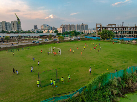 Aerial view of a soccer field with players practicing on green grass, surrounded by urban buildings and construction sites in Gurgaon, Haryana, India.