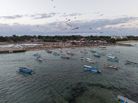 Aerial view of Mertasari Beach during a kite festival with numerous traditional boats anchored in the clear water and a large crowd gathered on the shore in Denpasar Selatan, Bali, Indonesia.