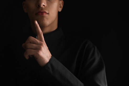 Priest in cassock pointing upwards on black background, closeup. Space for text