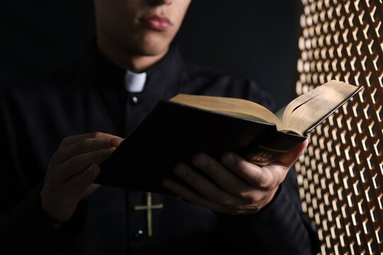 Priest with Holy Bible praying near partition in confessional booth, closeup