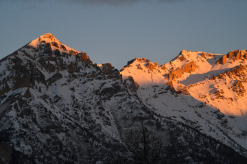 Obraz premium Warm sunrise lighting the top of snowy mountain peaks in the Piedmont region, Italy. Stunning morning alpenglow taken from Sauze D'oulx alpine ski resort. Dramatic snow-capped winter mountains at dawn
