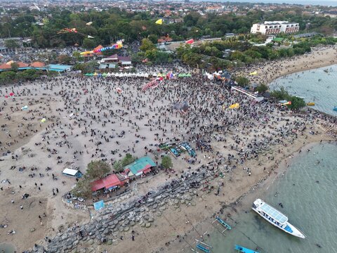Aerial view of Mertasari Beach crowded with people during a kite festival with colorful kites, boats moored in the water, and trees in Denpasar Selatan, Bali, Indonesia.
