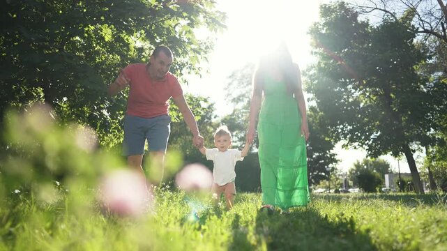 Child walk with mom father park. Parents hold child hand guiding first step. Family enjoys outdoor walk together. Child learning to walk with mom father support. Happy moments family walk first step.