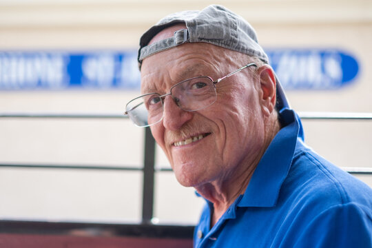 Senior adult man with a mustache and eyeglasses smiling warmly, wearing a blue polo shirt and a grey baseball cap, expressing a positive and happy demeanor
