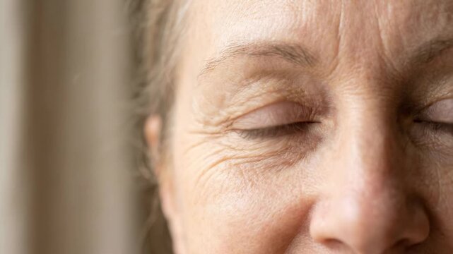 Tight eye level close up showing right eye and adjacent facial area of mature person with brown green iris, steady open gaze, visible skin texture, subtle wrinkles, soft catchlight, and natural calm