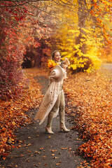 Fototapeta premium Woman walks along a path covered with autumn leaves while holding a dog and wearing a large hat with yellow leaves
