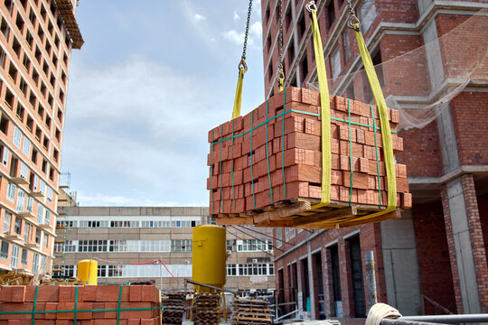 Crane lifting pallet of red clay bricks beside unfinished brick building at active construction site with workers, scaffolding, daylight sky and urban development context
