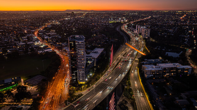 Aerial view of the Citylink Sound Tube illuminated at sunset with traffic flowing along the highway, surrounded by urban buildings and city lights in Flemington, Victoria, Australia.