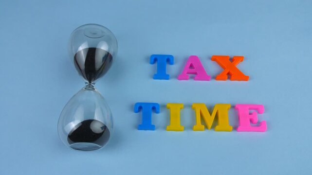 Zoom in shot of colorful 'Tax Time' words beautifully arranged on a blue platform. Closeup shot of a sand clock and words "Tax Time" composed with plastic letters - business/finance concept