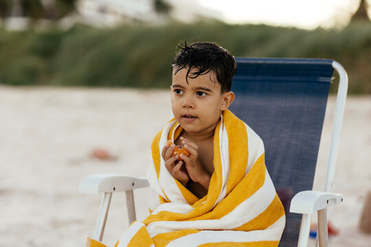 A young boy wrapped in a yellow striped beach towel sits on a folding chair at the seaside, holding a peeled orange and looking thoughtful after a swim.