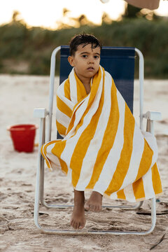 Pensive Young Boy Wrapped in Yellow Striped Towel Sitting on Beach Chair at Sunset