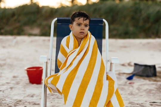 Pensive Young Boy Wrapped in Yellow Striped Towel Sitting on Beach Chair at Sunset