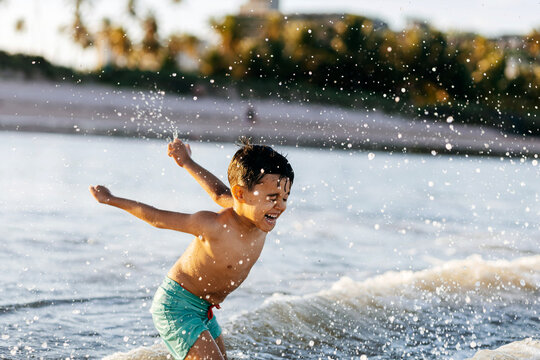 A smiling boy plays in shallow ocean waves, splashing water and laughing on a sunny tropical beach during a carefree summer vacation.