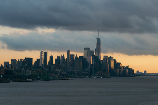 View of the Manhattan skyline featuring One World Trade Center under a dramatic cloudy sky at sunset, with the Hudson River in the foreground New York, New York, United States.