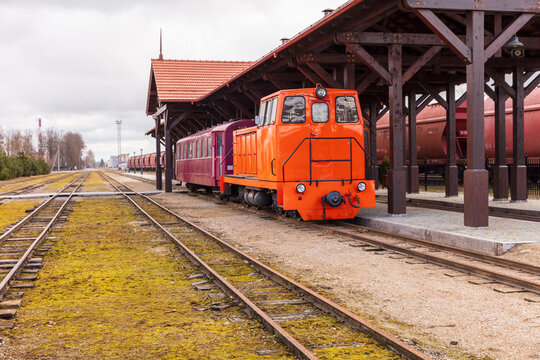 View of the historic Aukstaitija narrow gauge railway with its orange locomotive and purple carriage parked under a wooden station platform in Panevezys, Panevezio apskritis, Lithuania.