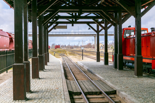 View of the Aukstaitija narrow gauge railway station platform with a red locomotive parked on the tracks under a wooden shelter in Panevezys, Panevezio apskritis, Lithuania.