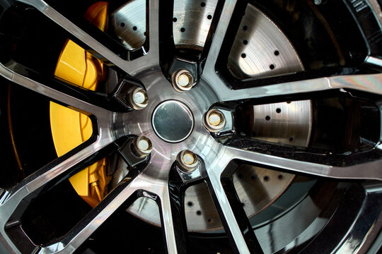 Detailed closeup of dark multi spoke alloy wheel on performance car showing drilled brake rotor, bright yellow brake caliper, exposed lug nuts, metallic rim, and precision mechanical surface textures
